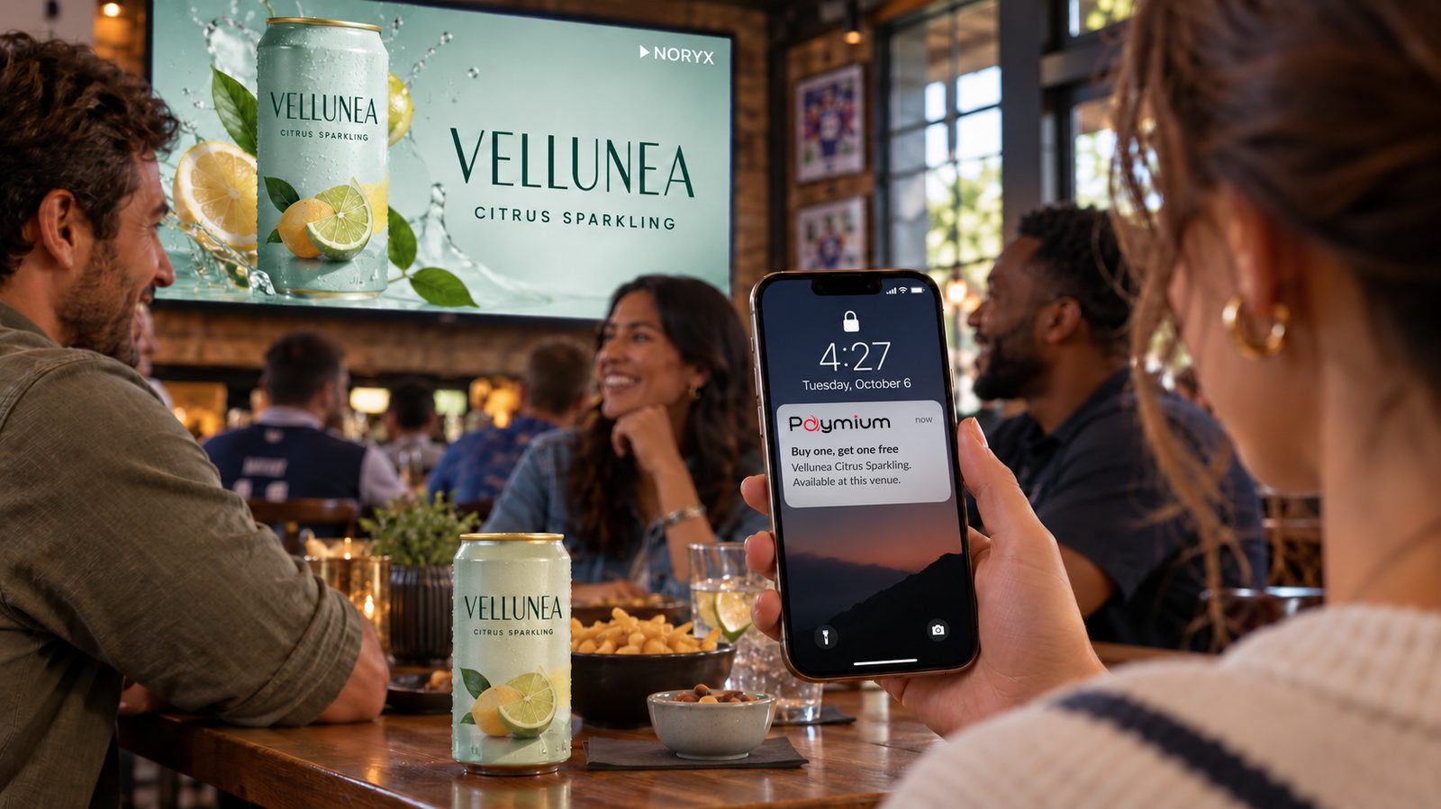 A drink commercial plays on the bar screen while a guest holds a locked phone showing a Paymium notification: buy one, get one free on the same drink at this venue.